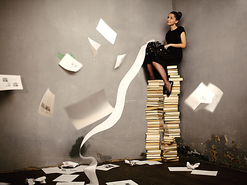 woman sitting on high stack of books with a typewriter, looks like she is thining and papers are flyign around,get paid to write articles, can i get paid to write, get paid to write 2022, newsbreak creator login, freelance writing jobs online, freelance writing jobs from home, freelance writing sites